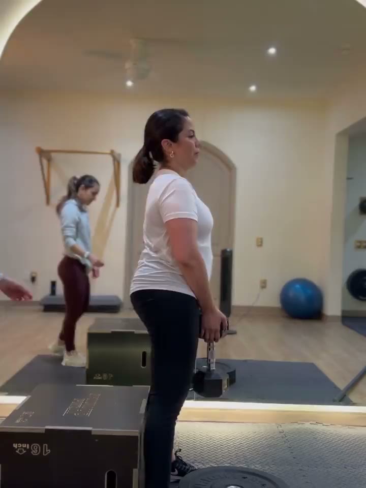Una clienta levantando una mancuerna desde una plataforma mientras la entrenadora la acompaña